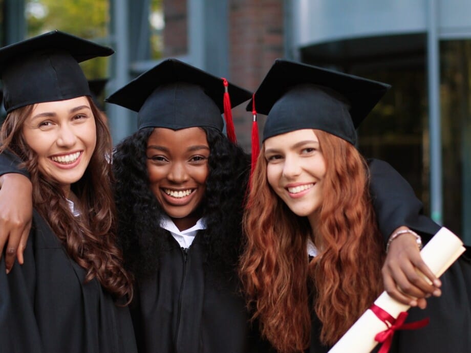 Happy group of mature students on graduation day embracing with each other. Three best girl friends in academic gowns and caps hugging in front of the camera