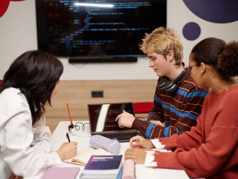 Diverse group of college students studying IT together at table in school library and using laptop with code lines on screen